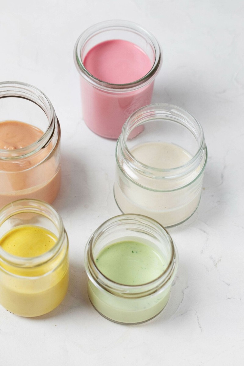 Five glass mason jars rest on a white surface. Each is filled with a different color of creamy tahini dressing.